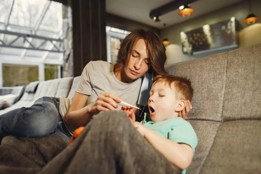 Mother taking sons temperature while sitting on sofa in apartment