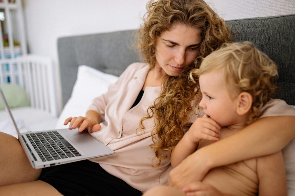 A mother with curly blonde hair sits with her toddler son on a bed, using a laptop.