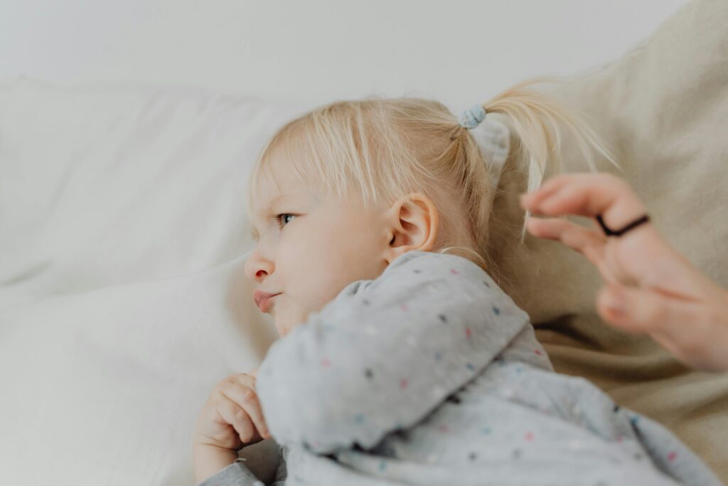 Cute blonde child with ponytail sulking on a soft bed indoors.